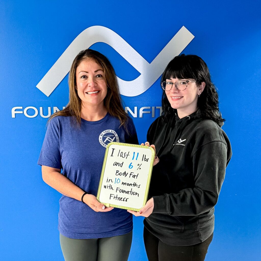 A Foundation Fitness client holding a sign with the weight and body fat she lost, standing with her trainer